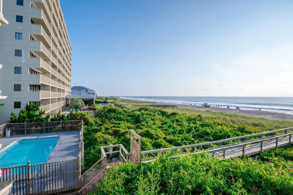 An inviting oceanfront pool sits in the foreground, surrounded by lush green landscaping and a private boardwalk leading directly to the sandy beach. The tranquil scene is framed by sunrise light, with clear views of the pool, poolside loungers, and the sparkling ocean beyond.