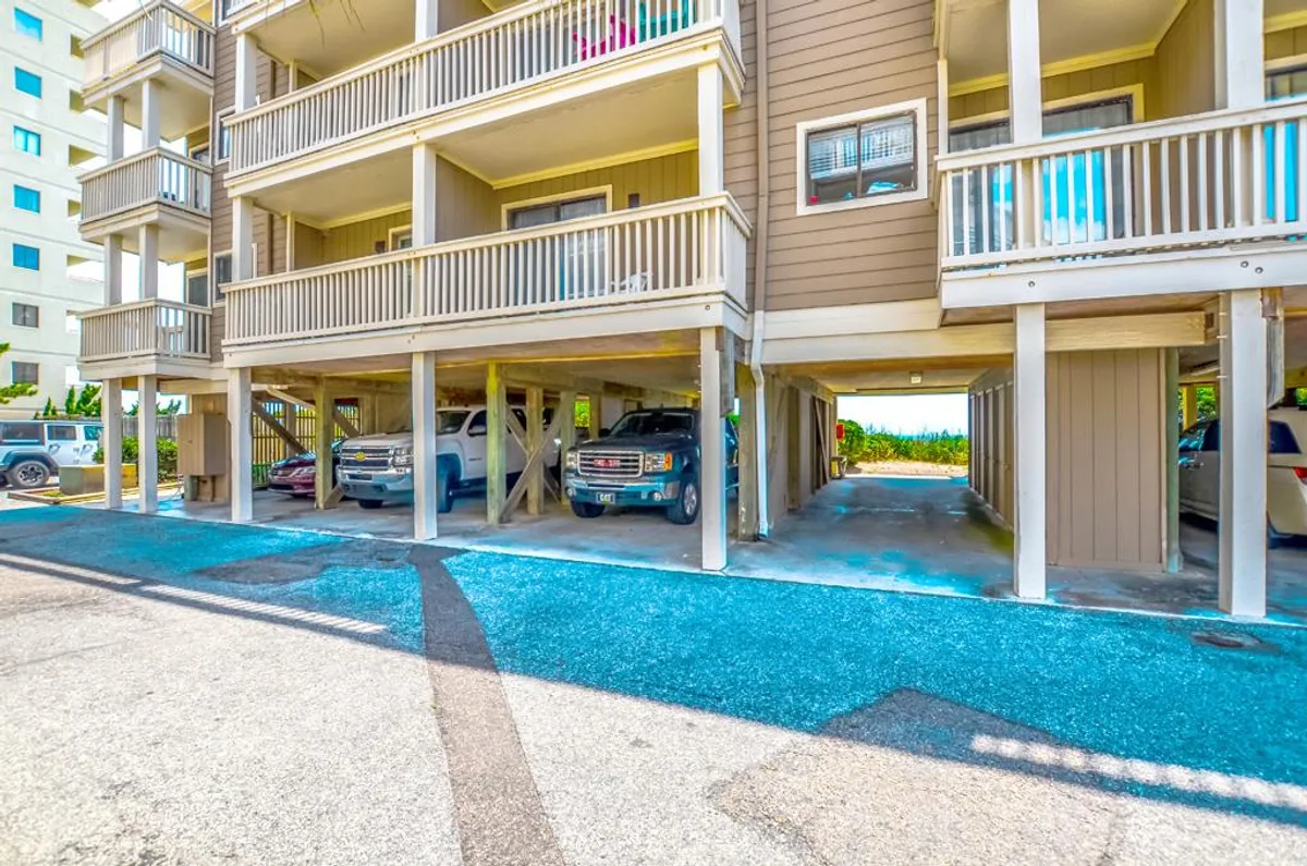 The covered parking area underneath the condo building is visible, with cars parked and some open spaces. The structural design of the building and the blue gravel driveway are prominent, offering a sense of convenience and security.