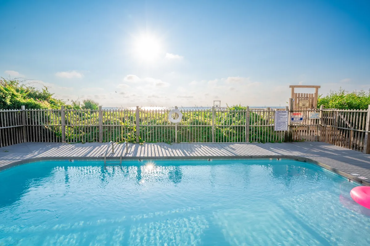 This cheerful exterior shot centers on the sparkling swimming pool, with sunlight dancing on the water. The fenced area and a visible playground structure in the background suggest a safe, family-friendly environment perfect for swimming, sunbathing, and kids’ play.