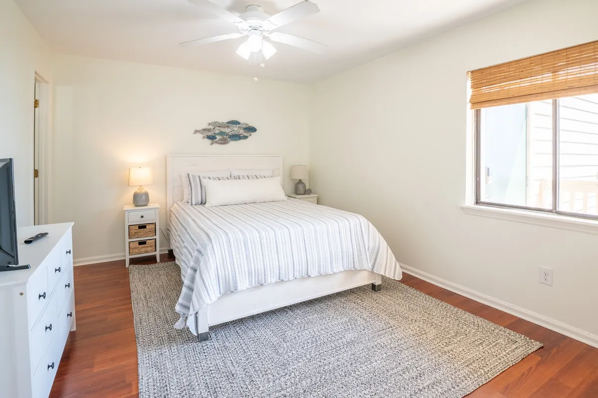 A serene bedroom featuring a queen bed topped with plush linens, a nightstand with a gentle lamp, and a window allowing natural sunlight to stream in. The calm decor and soft area rug add to the tranquil, restful atmosphere. A TV is mounted on the wall, complementing the room's modern, comfort-oriented design.