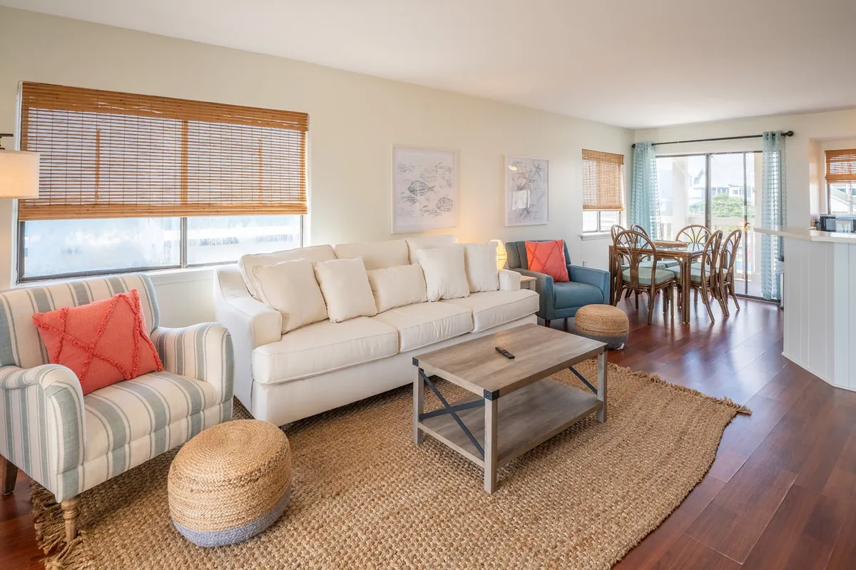 This interior scene displays a cozy living and dining area, with a dining table, woven rug, and ample natural light. The table is set for group gatherings and is adjacent to a lounge space, emphasizing warmth and connection.