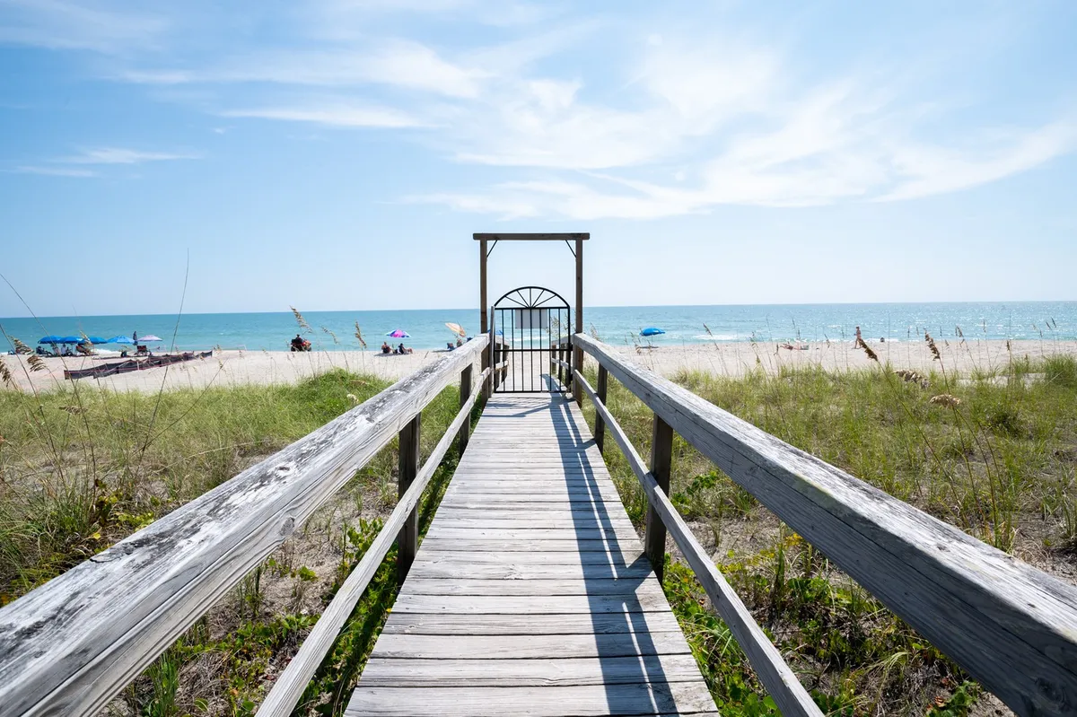 This image features a wooden walkway leading from the property towards a beach gate, with a sweeping view of the ocean just beyond. The scene is inviting and highlights the property's direct and private access to the beach, framed by gentle coastal greenery and soft sunlight.