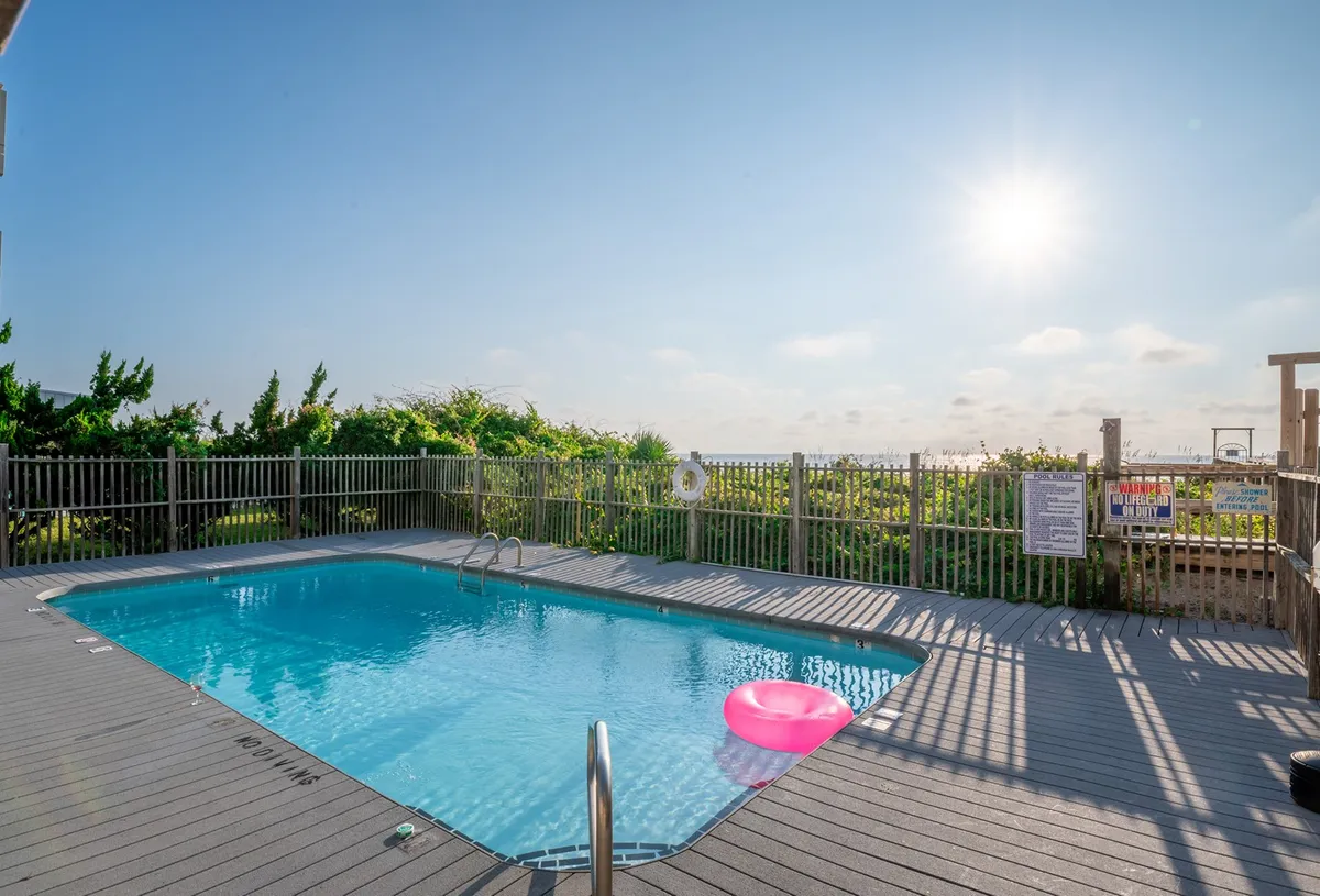 This exterior shot captures a sunlit swimming pool surrounded by a fence and landscaping, with a pink float bobbing on the water. The inviting scene radiates energy, suggesting a lively spot to unwind after a day of adventure. The pool area is clearly a hub for socializing, sunbathing, and enjoying waterfront relaxation, with sunlight casting a warm glow across the scene.