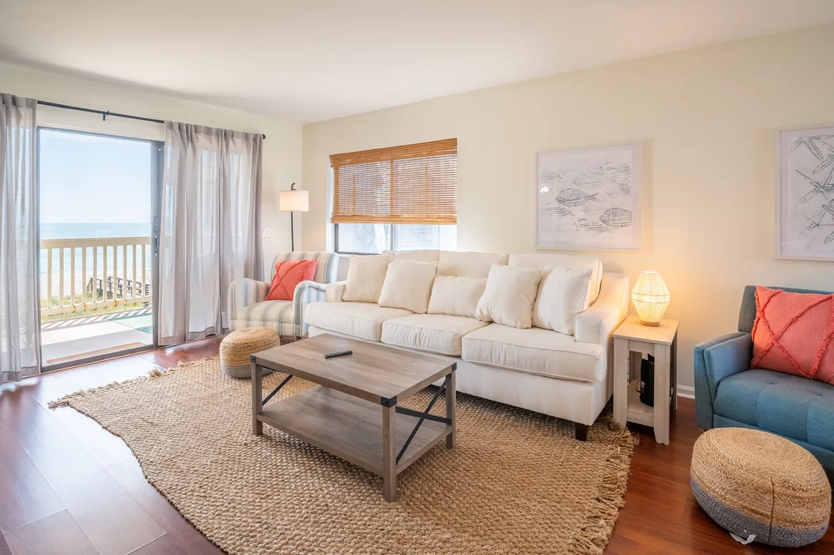 This image captures a bright, open living room with a large white sofa, a wooden coffee table, colorful accent chairs, and—most importantly—an expansive ocean view visible through the sliding glass door. Natural light floods the space, highlighting the new, plush furnishings and the seamless flow from the interior to the porch.