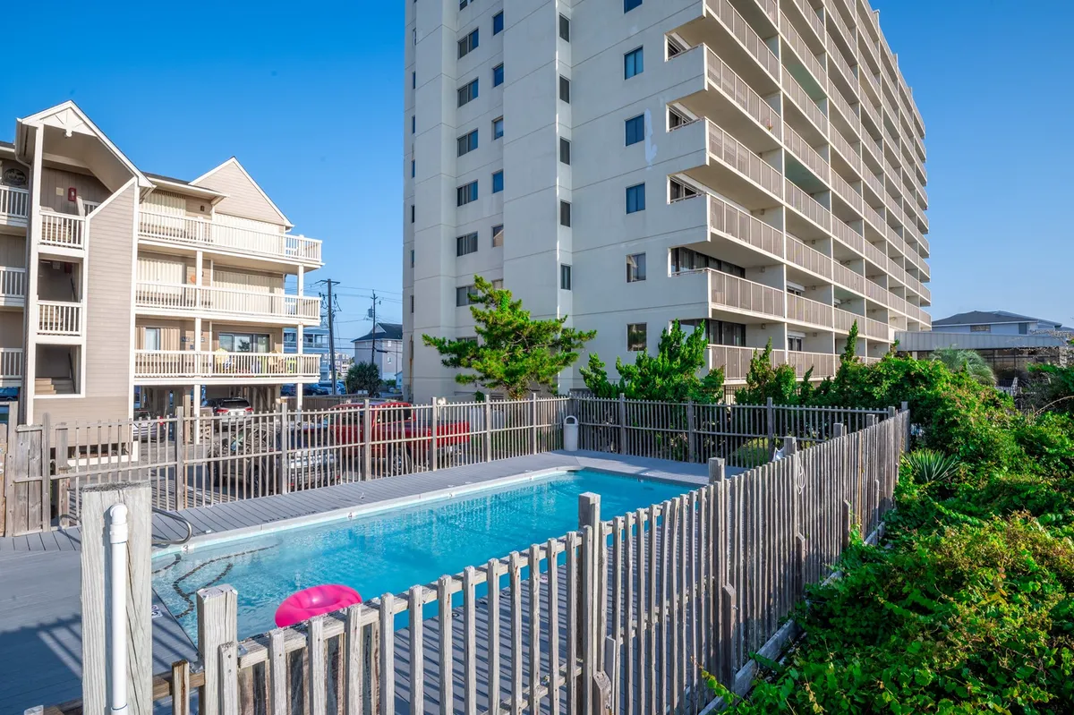 An inviting outdoor swimming pool is framed by fencing and surrounded by nearby buildings, offering a communal area for relaxing, socializing, or sunbathing during a sunny day. The scene encourages outdoor gathering and fun, connecting guests to the larger community and summer atmosphere.
