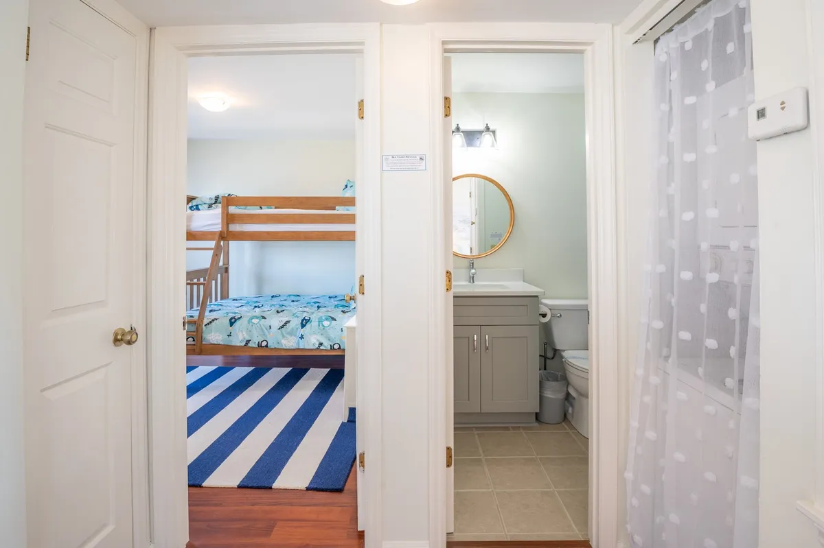 A bright, family-friendly bedroom with a twin-over-full bunk bed, a colorful striped rug, and a bathroom vanity visible just beyond the bed. The room is thoughtfully arranged, making it inviting for both children and adults. The decor is fresh and playful, contributing to a welcoming sanctuary feel.
